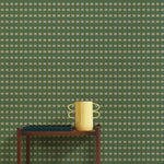 A wallpapered wall in a gridded check pattern in greens with a wooden cart with a yellow vase, green scalloped tray and a stack of 3 books. 