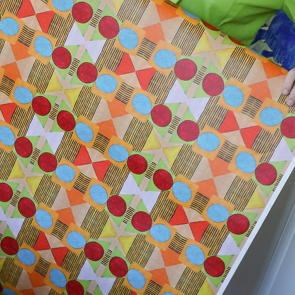 Detail of a woman holding a wallpaper with a collaged circle, square and diamond stripe wallpaper in red, orange, green and pink. 