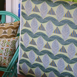 Detail of a woman holding a wallpaper with a collaged leaf pattern in blue, green, yellow and grey, next to a turquoise chair with patterned throw pillows. 