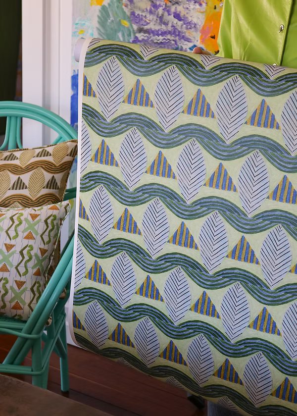 Detail of a woman holding a wallpaper with a collaged leaf pattern in blue, green, yellow and grey, next to a turquoise chair with patterned throw pillows. 