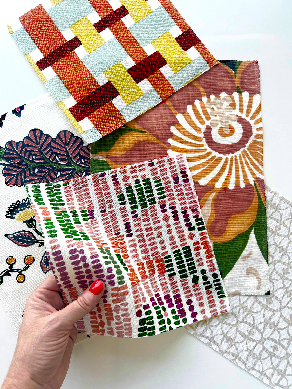 A hand with red nail polish holds a square fabric swatch featuring a pink, green, and orange dotted pattern. In the background, several other patterned fabric swatches are arranged on a white surface, including a geometric orange, yellow, and blue design, a large floral print in brown, orange, and green, and a white pattern with small orange berries.