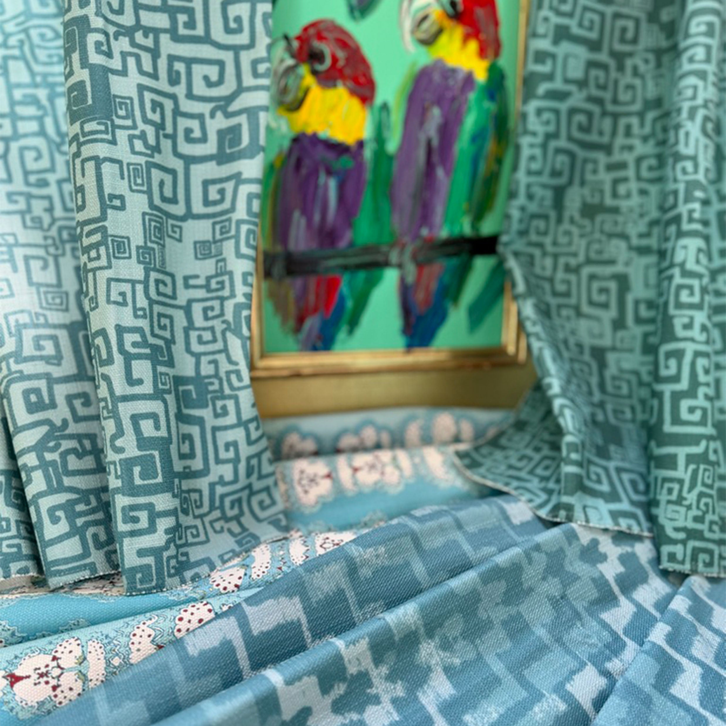 Draped swatches of blue patterned fabrics against a colorful painting of parrots. 