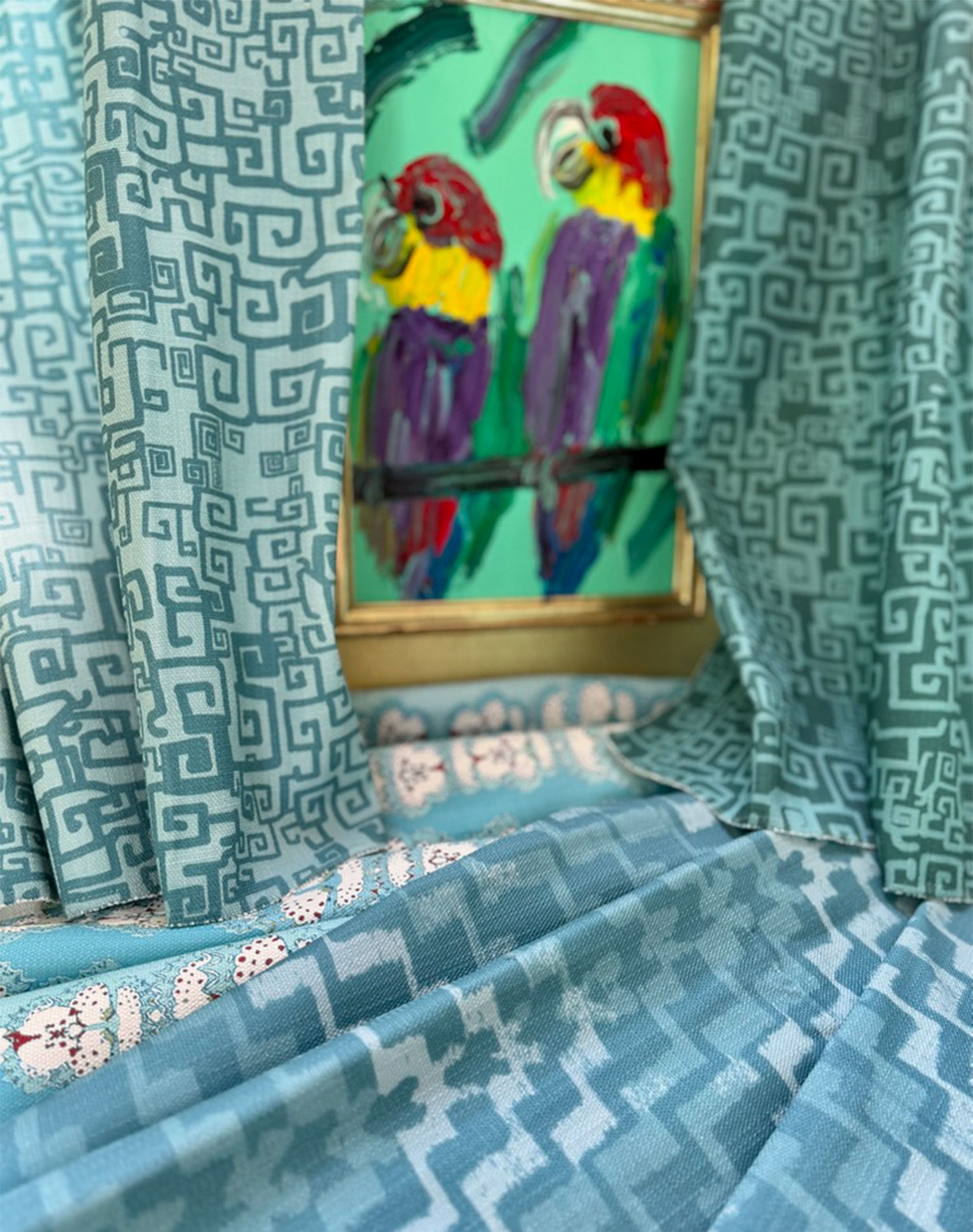 Draped swatches of blue patterned fabrics against a colorful painting of parrots. 