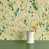 Vase of flowers stands in front of a wall papered in a painterly leaf print in shades of green, white and yellow.
