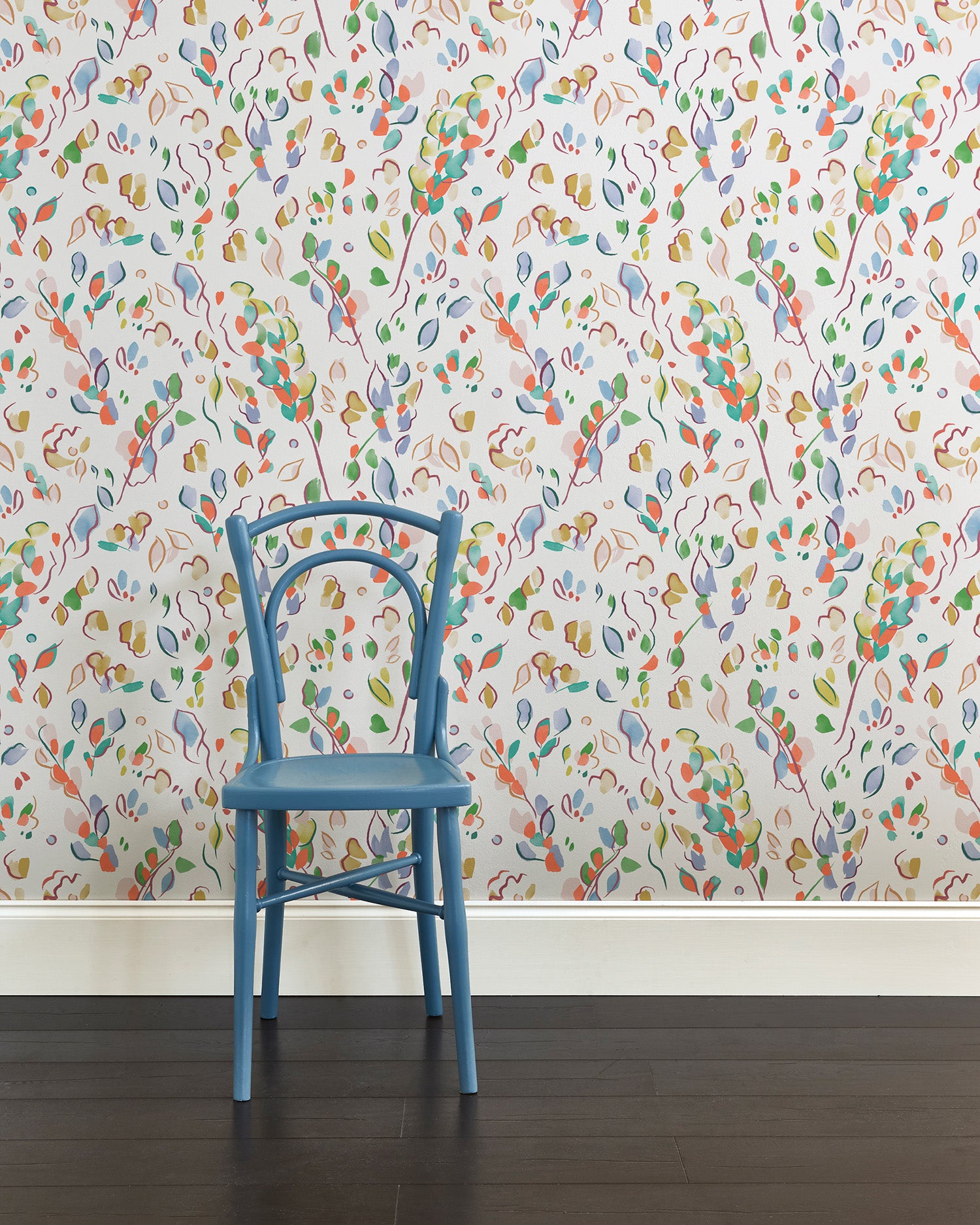 A blue chair stands in front of a wall papered in a painterly leaf print in a rainbow of shades on a white field.