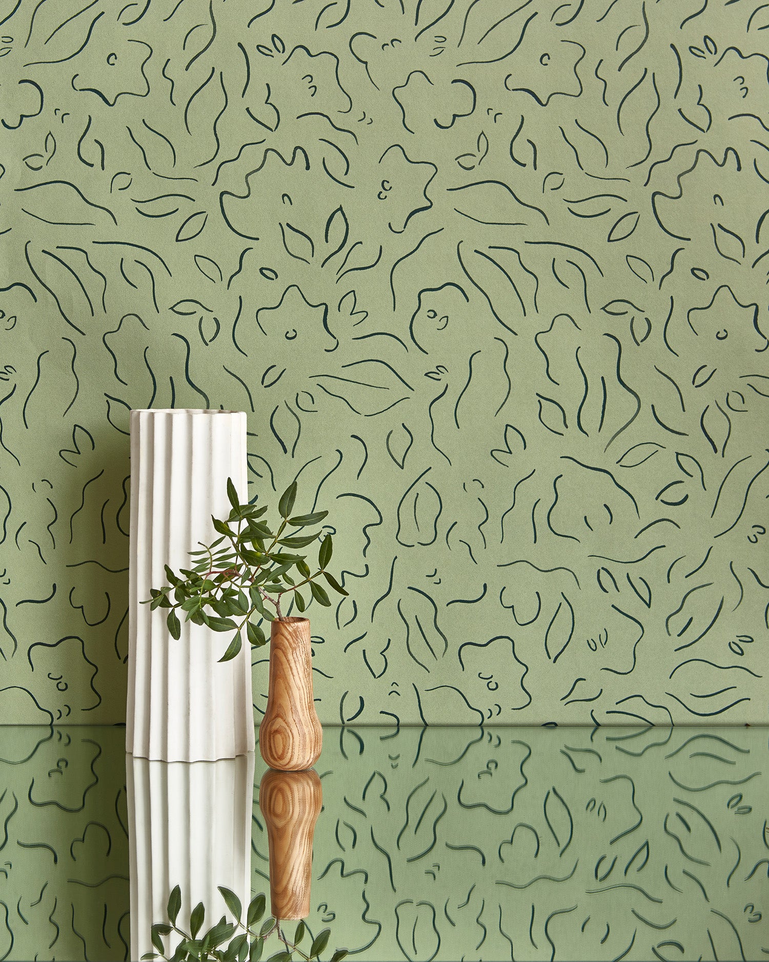 Two vases stand in front of a wall papered in a minimalist floral print in sage on a green field.