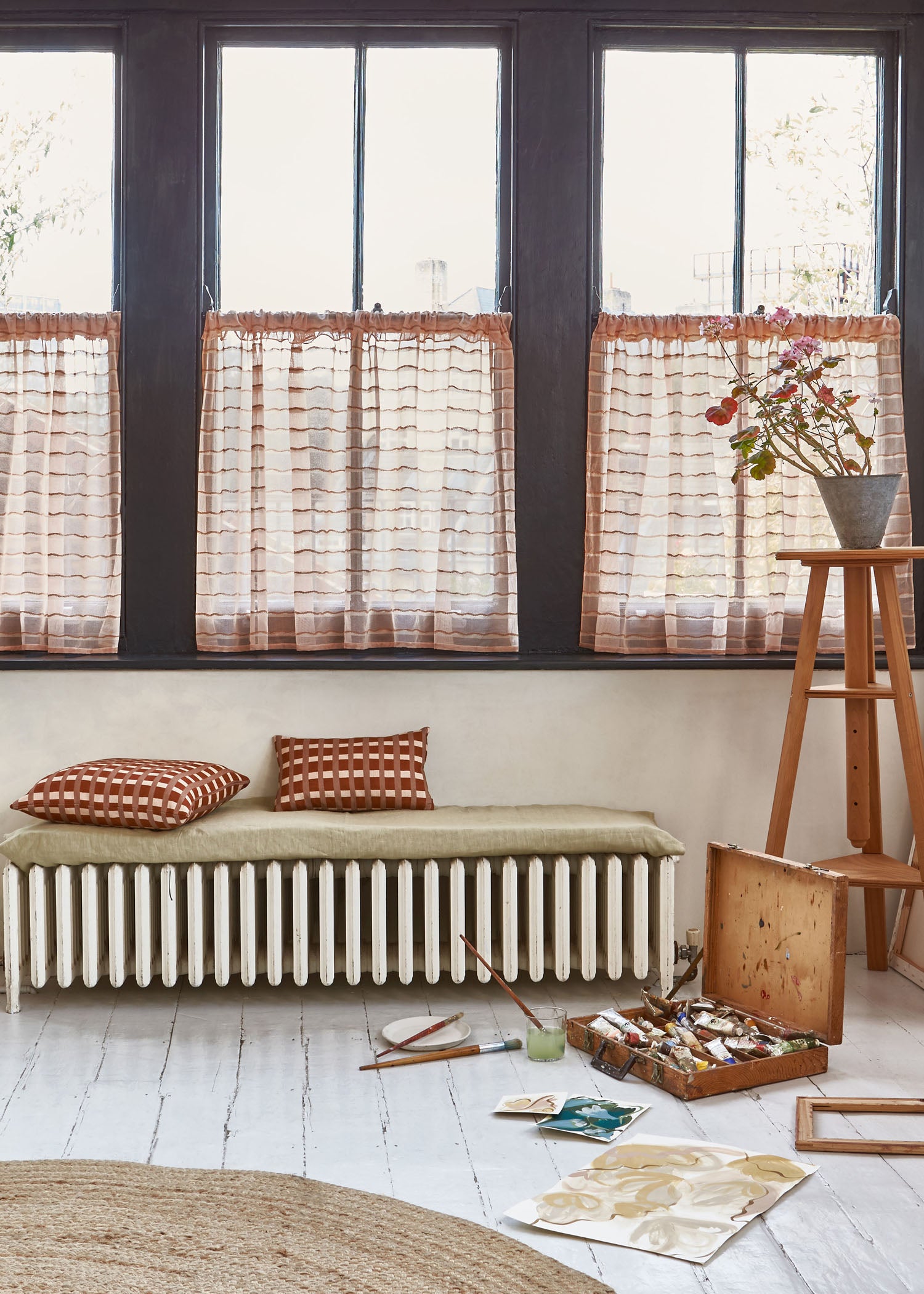 An artists studio interior with a paint kit, brushes and artwork scattered on the floor against a wall of windows with  sheer blush colored curtains in blush pink.