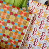 Cropped image of two figured holding large pieces of geometric wallpapers in bright orange, red, yellow and white. 
