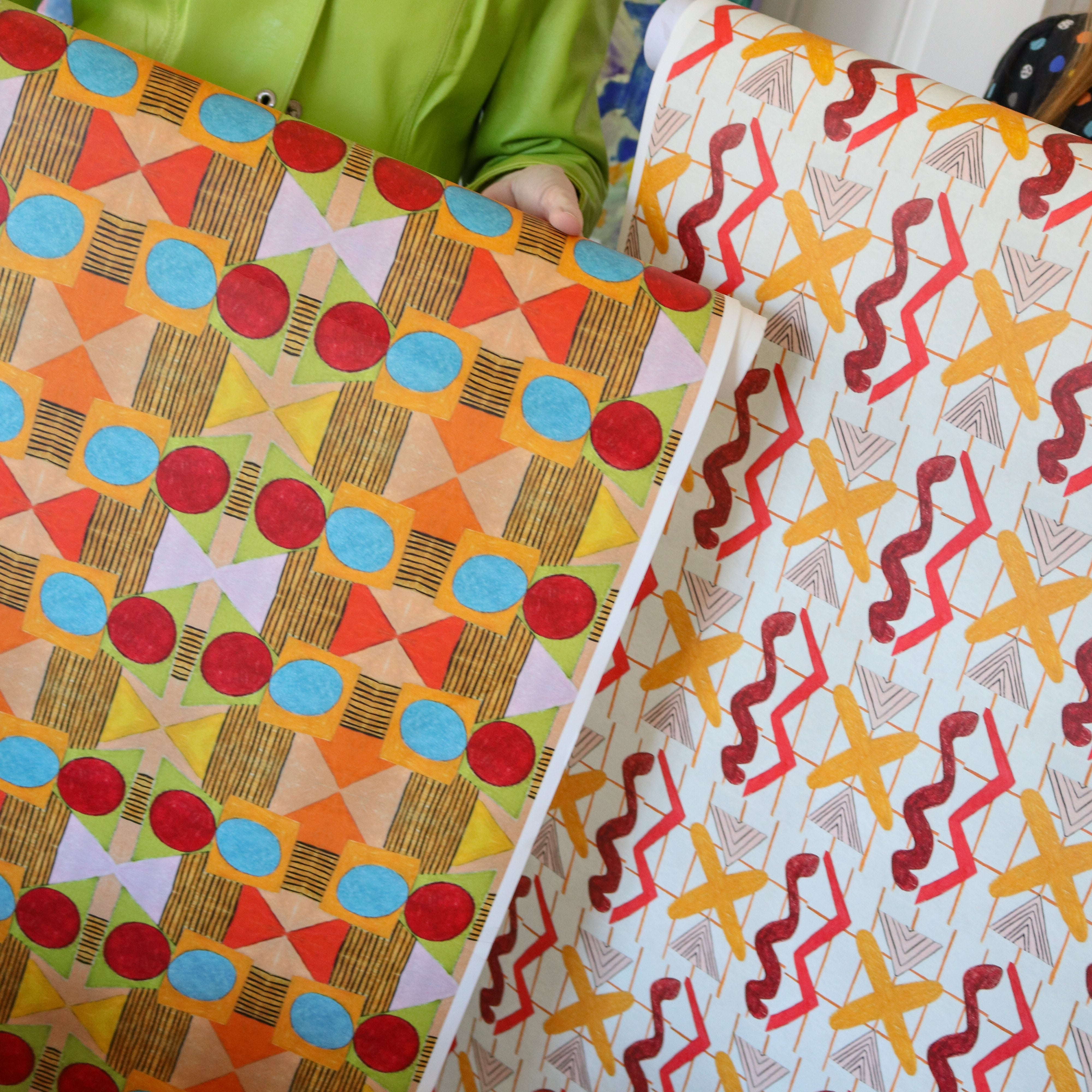 Cropped image of two figured holding large pieces of geometric wallpapers in bright orange, red, yellow and white. 