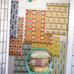 Interior setting looking through a doorway to with a turquoise rattan chair with printed throw pillows against a white wainscoted wall with a collage of geometric patterned wallpapers in bright colors. 