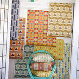 Interior setting looking through a doorway to with a turquoise rattan chair with printed throw pillows against a white wainscoted wall with a collage of geometric patterned wallpapers in bright colors. 