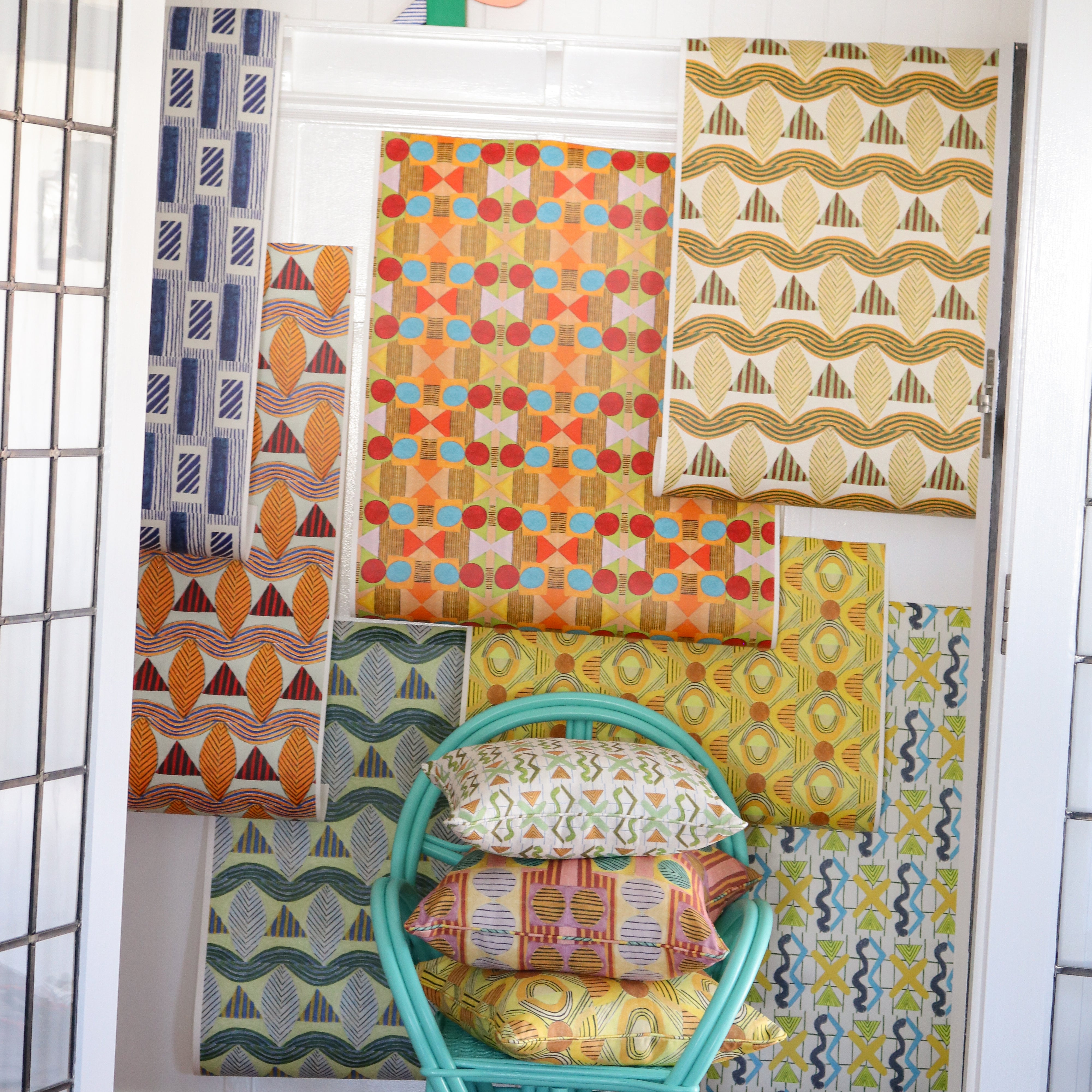 Interior setting looking through a doorway to with a turquoise rattan chair with printed throw pillows against a white wainscoted wall with a collage of geometric patterned wallpapers in bright colors. 
