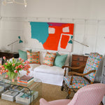 living room scene with pink upholstered side chairs, plexiglass coffee table with stacked magazines below and on top with a vase of pink tulips, White couch with bold patterned throw pillows against a white wall with decorative paneling and a large abstract painting in teal, white and red. 