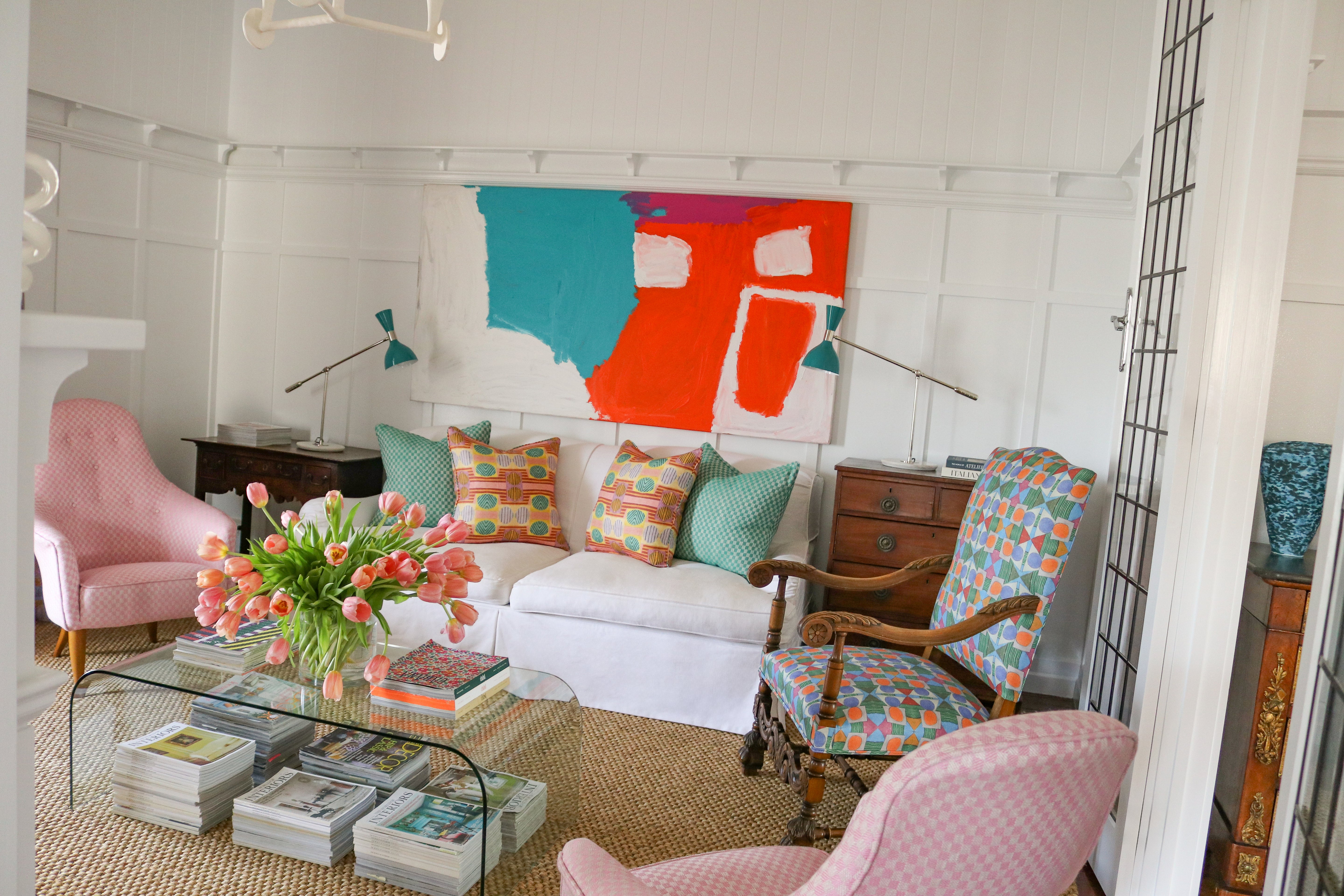 living room scene with pink upholstered side chairs, plexiglass coffee table with stacked magazines below and on top with a vase of pink tulips, White couch with bold patterned throw pillows against a white wall with decorative paneling and a large abstract painting in teal, white and red. 
