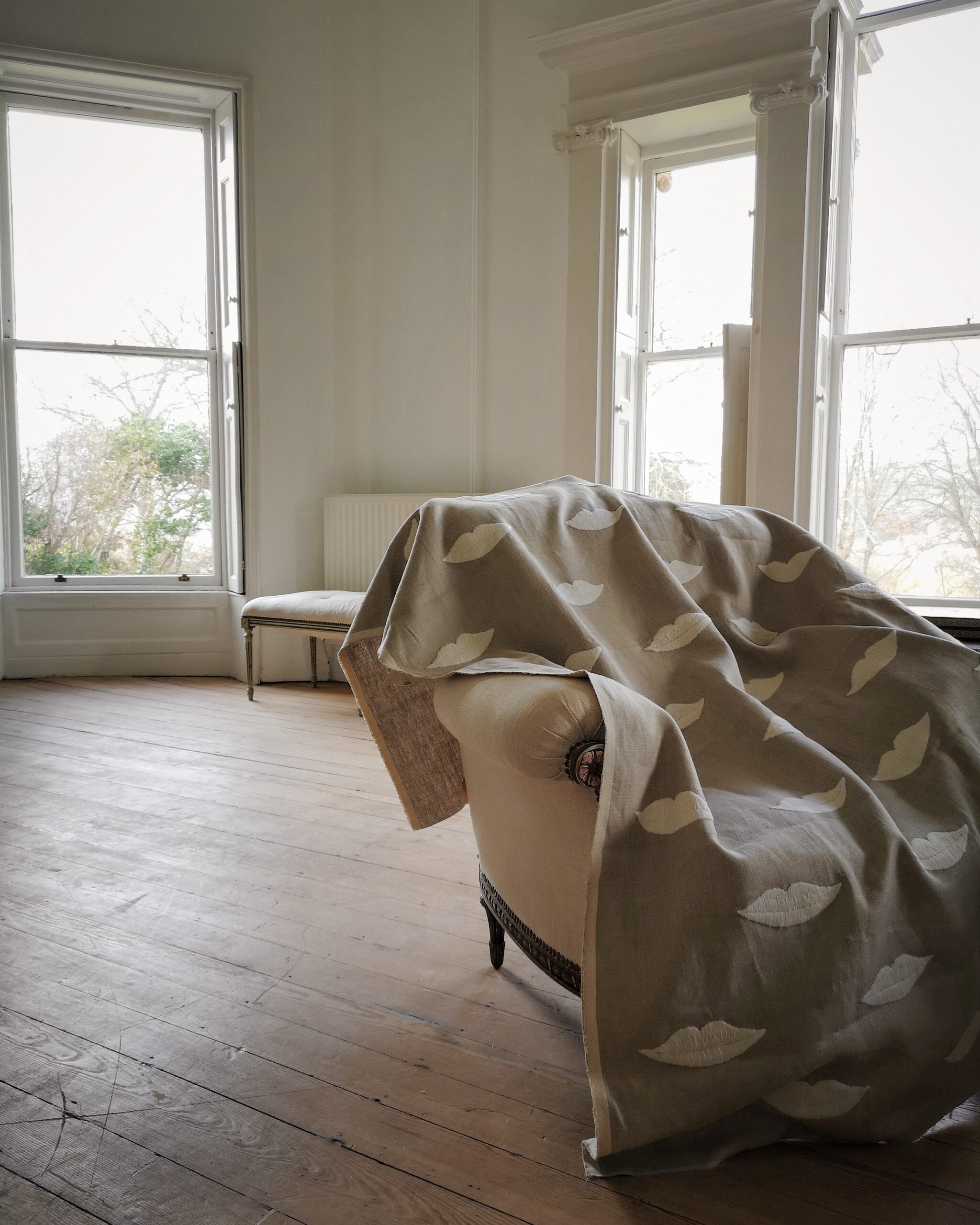 Room with a embroidered linen panel with white lips draped over a chair, large windows allowing natural light.