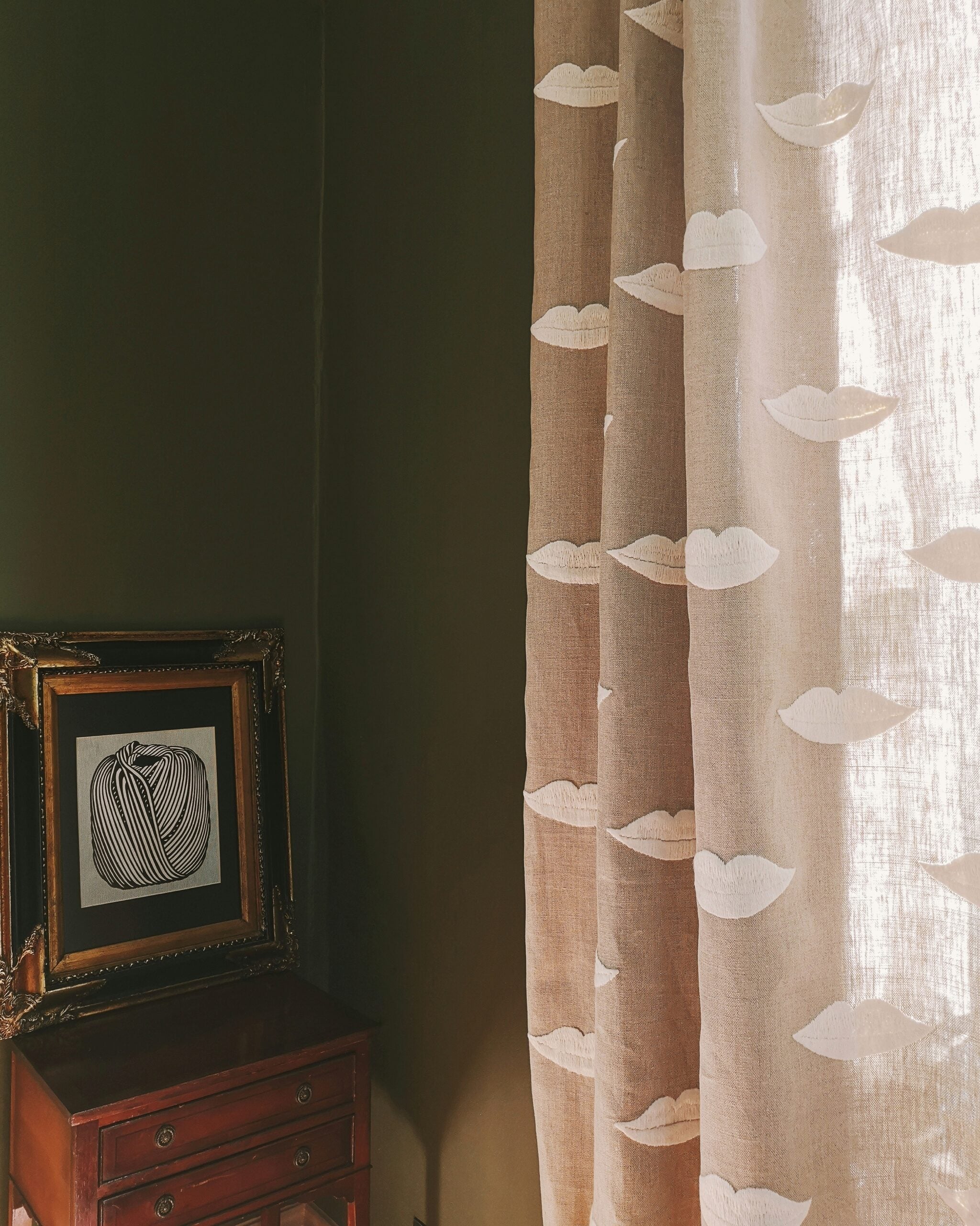 Linen curtain with white lip embroidery in a room with a wooden table and framed artwork.