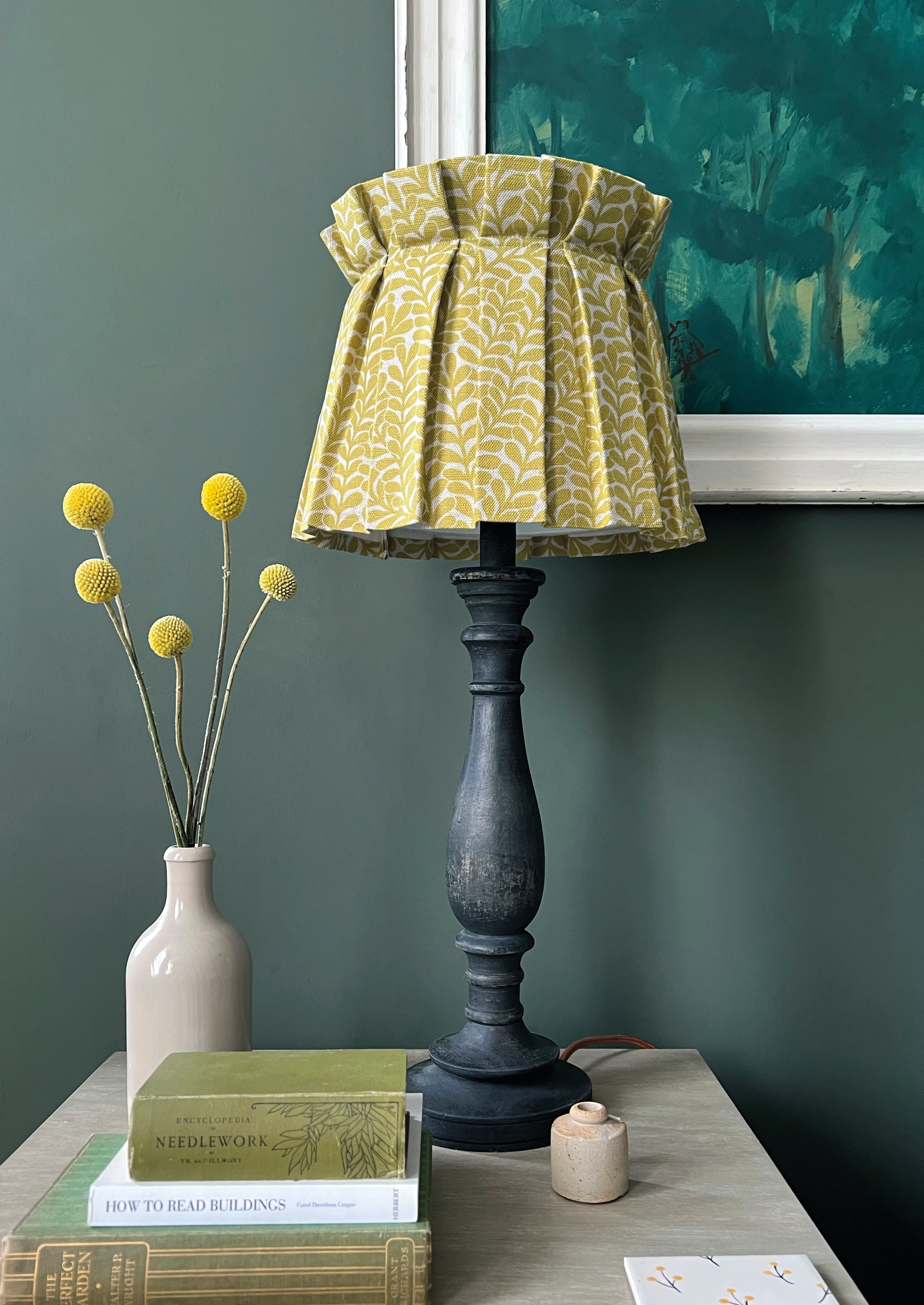 An end table with flowers, a stack of books and a lamp with a shade upholstered in a scrolling stem and leaf print.