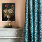 A dresser and floral painting stand next to a curtain in a scrolling stem and leaf print in turquoise on a white field.