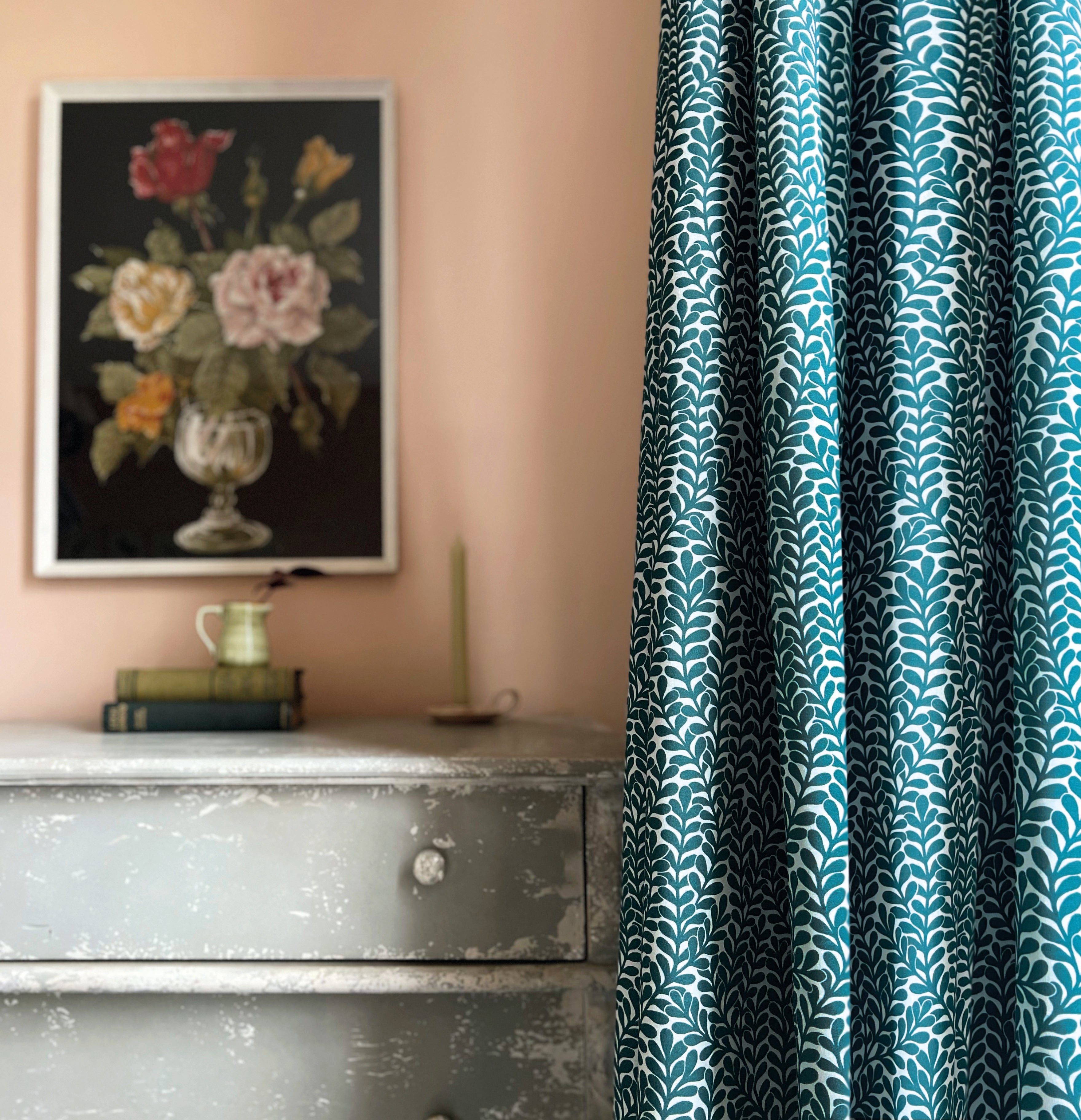 A dresser and floral painting stand next to a curtain in a scrolling stem and leaf print in turquoise on a white field.