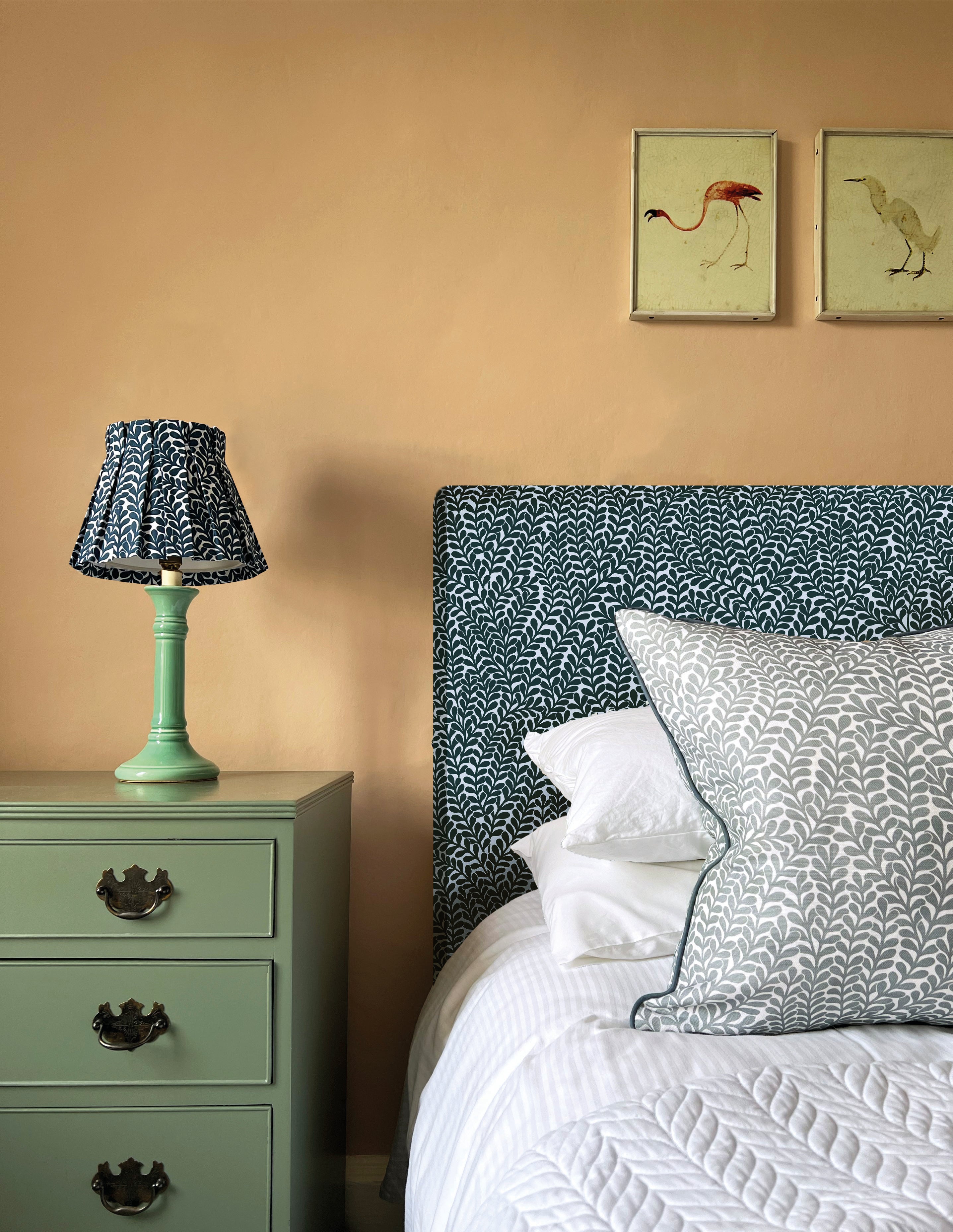 A cozy bedroom tableau with a headboard, lamp and throw pillows all upholstered in the same scrolling stem and leaf print.