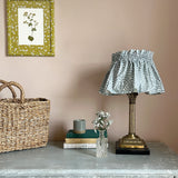 A dresser topped with a basket, books, and a lamp with a shade upholstered in a scrolling stem and leaf print.
