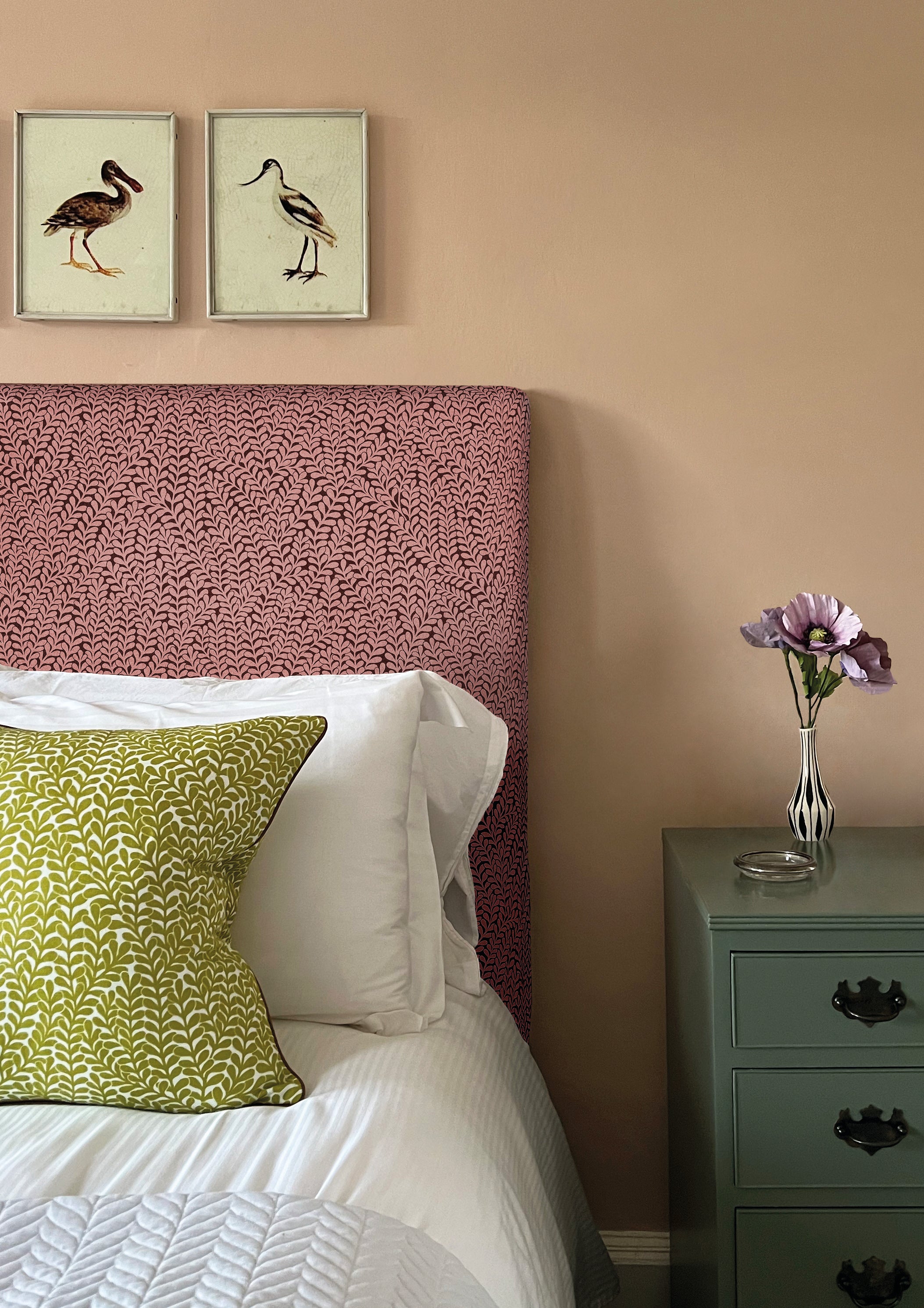 A cozy bedroom tableau with a headboard and throw pillows all upholstered in the same scrolling stem and leaf print.
