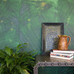 A large plant and an end table stand in front of a wall covered in multi-directional combed wallpaper in mottled green.