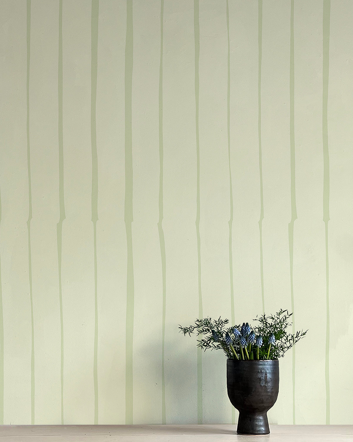 A vase of flowers stands in front of a wall covered in a playful irregular stripe print in light green on a cream field.