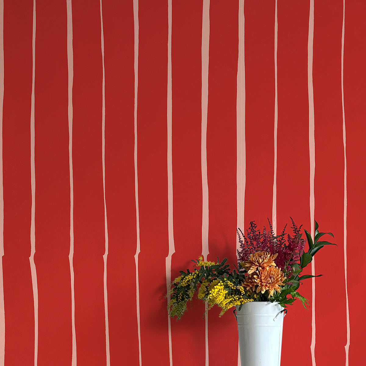 A vase of flowers stands in front of a wall covered in a playful irregular stripe print in cream on a red field.