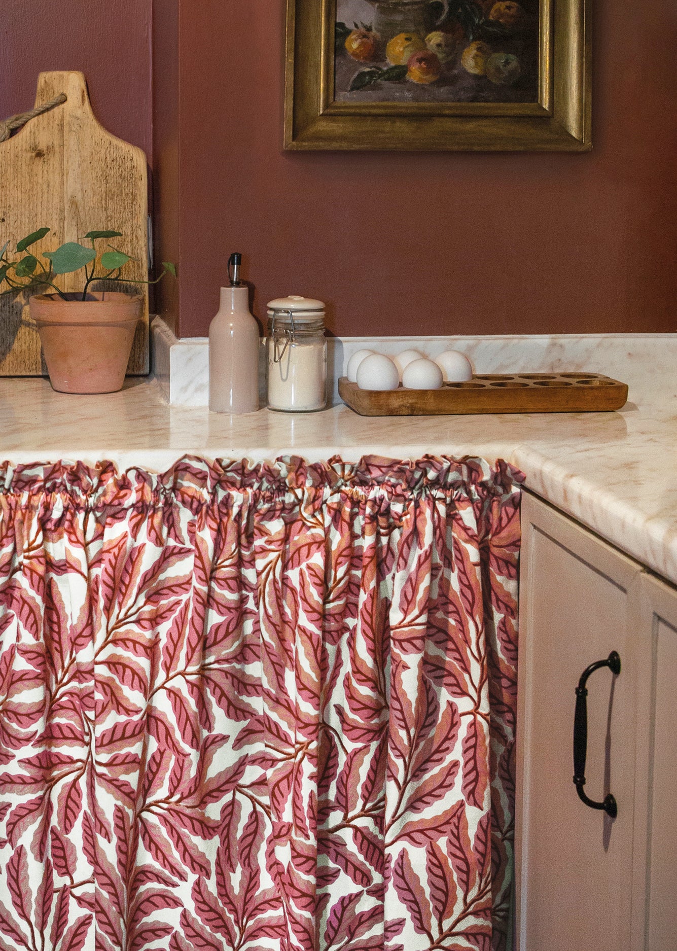 Kitchen with pink leaf-patterned curtain, countertop items, and a painting on the wall.