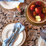 Dining table setting with plates, cutlery, and a bowl of fruit on a patterned tablecloth.
