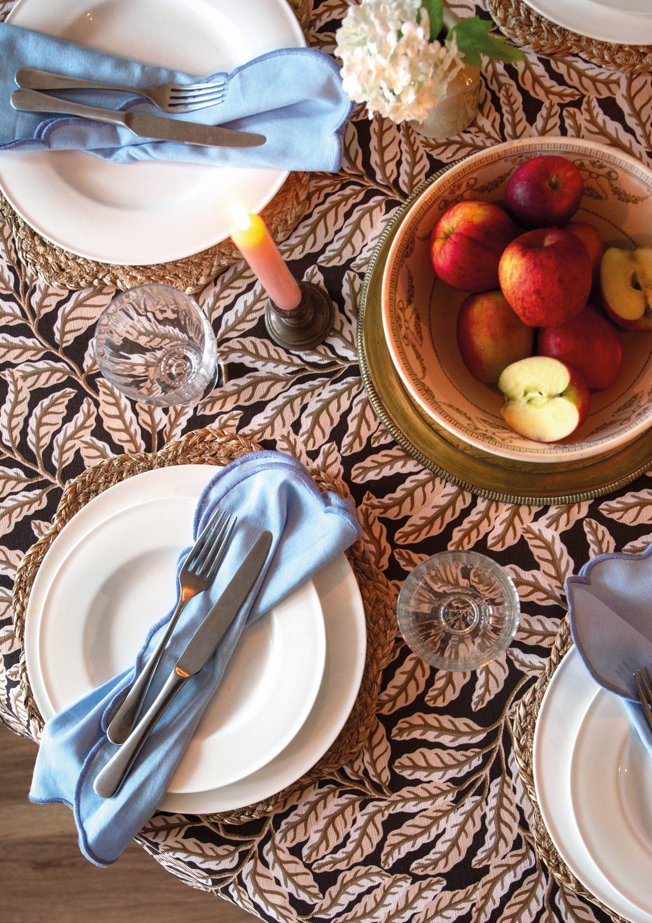 Dining table setting with plates, cutlery, and a bowl of fruit on a patterned tablecloth.