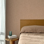 Bed with tan linens and wooden headboard, wooden side table  with white book and green glass, white floor length curtain against a watercolor swirling stripe wallpaper in tonal clay pink. 