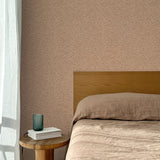 Bed with tan linens and wooden headboard, wooden side table  with white book and green glass, white floor length curtain against a watercolor swirling stripe wallpaper in tonal clay pink. 