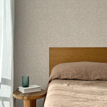 Bed with tan linens and wooden headboard, wooden side table  with white book and green glass, white floor length curtain against a watercolor swirling stripe wallpaper in tonal beige.