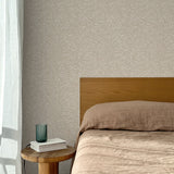 Bed with tan linens and wooden headboard, wooden side table  with white book and green glass, white floor length curtain against a watercolor swirling stripe wallpaper in tonal beige.
