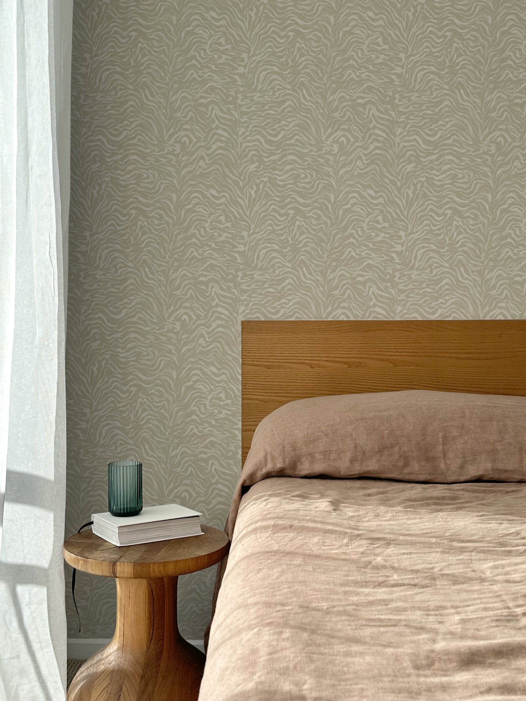 Bed with tan linens and wooden headboard, wooden side table  with white book and green glass, white floor length curtain against a watercolor swirling stripe wallpaper in tonal sage green.