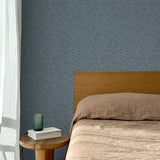 Bed with tan linens and wooden headboard, wooden side table  with white book and green glass, white floor length curtain against a watercolor swirling stripe wallpaper in tonal cool blue. 