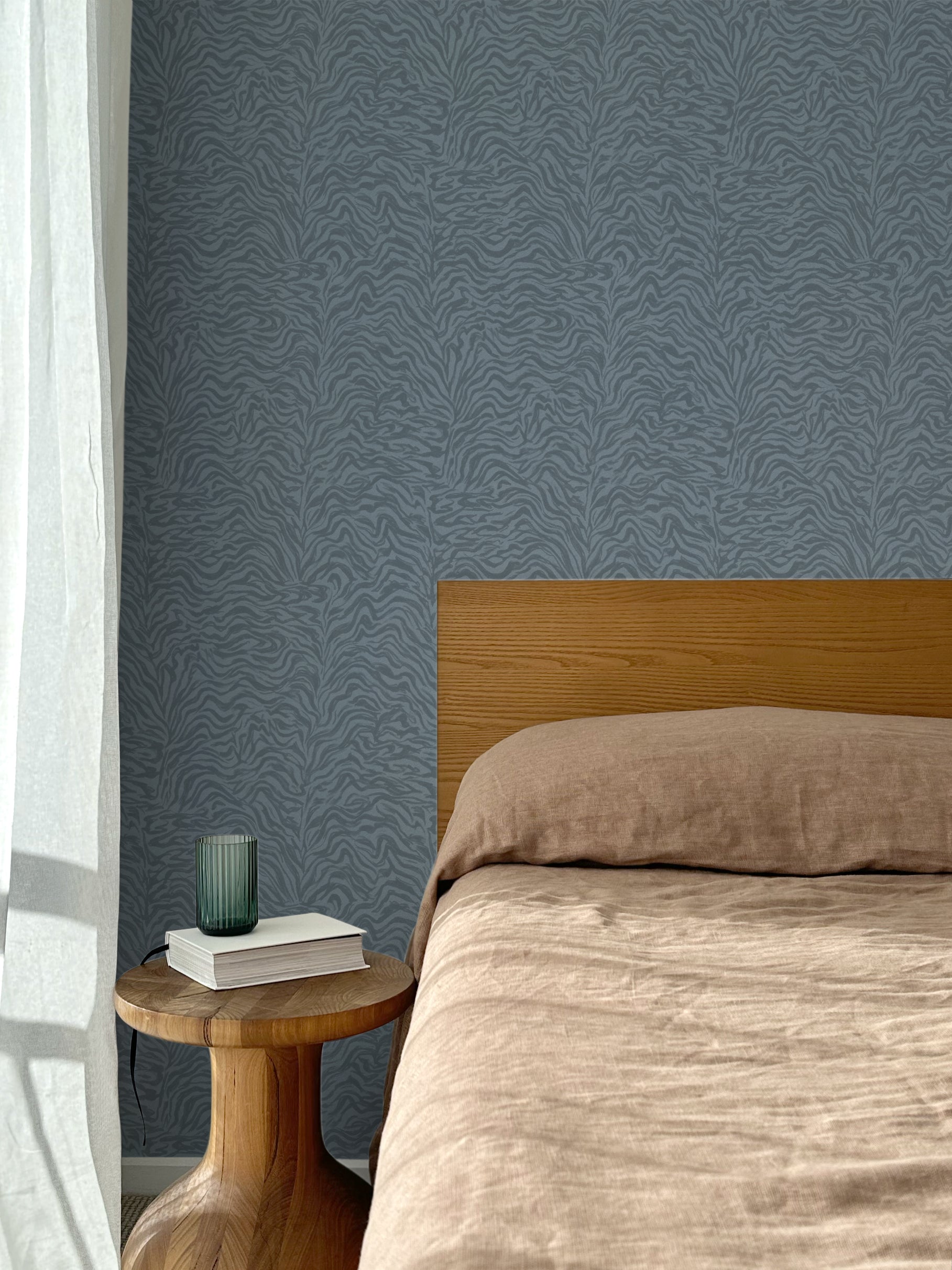 Bed with tan linens and wooden headboard, wooden side table  with white book and green glass, white floor length curtain against a watercolor swirling stripe wallpaper in tonal cool blue. 