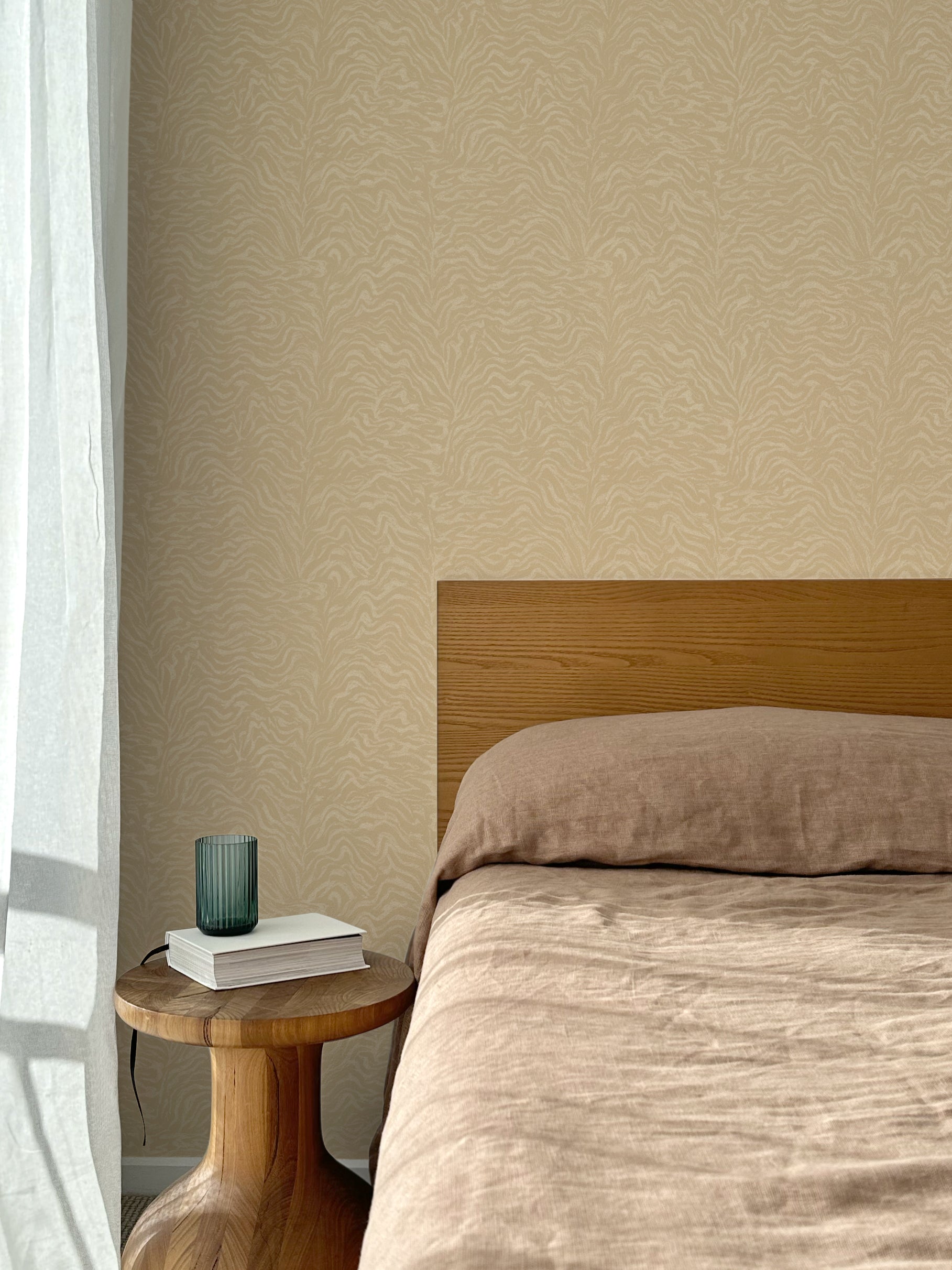 Bed with tan linens and wooden headboard, wooden side table  with white book and green glass, white floor length curtain against a watercolor swirling stripe wallpaper in tonal tan. 