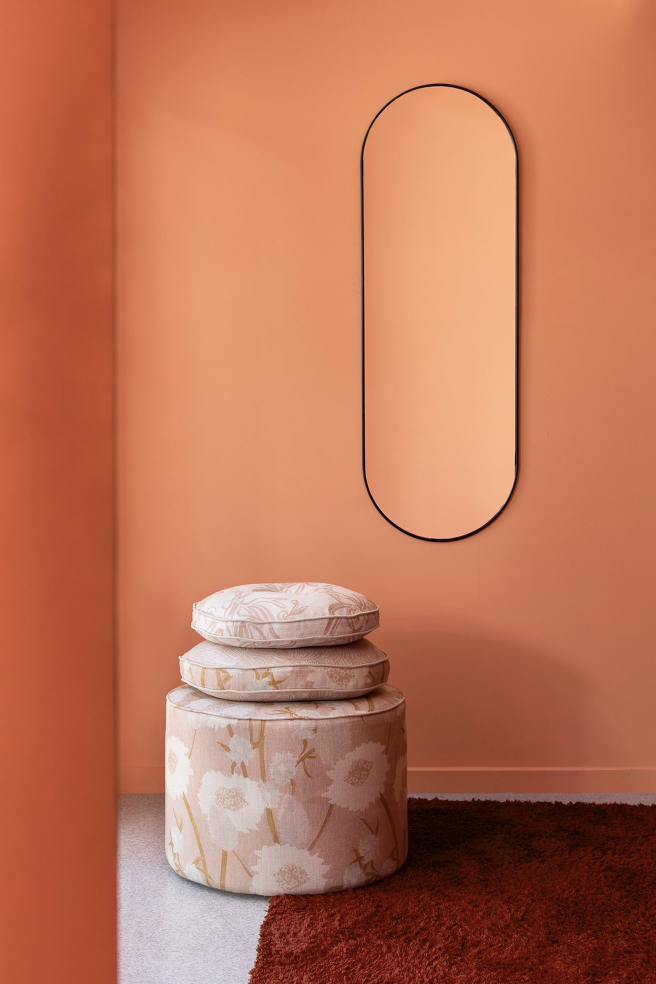 A pouf and stack of pillows, all in a playful floral print in light pink and tan, stand in front of a painted orange wall.