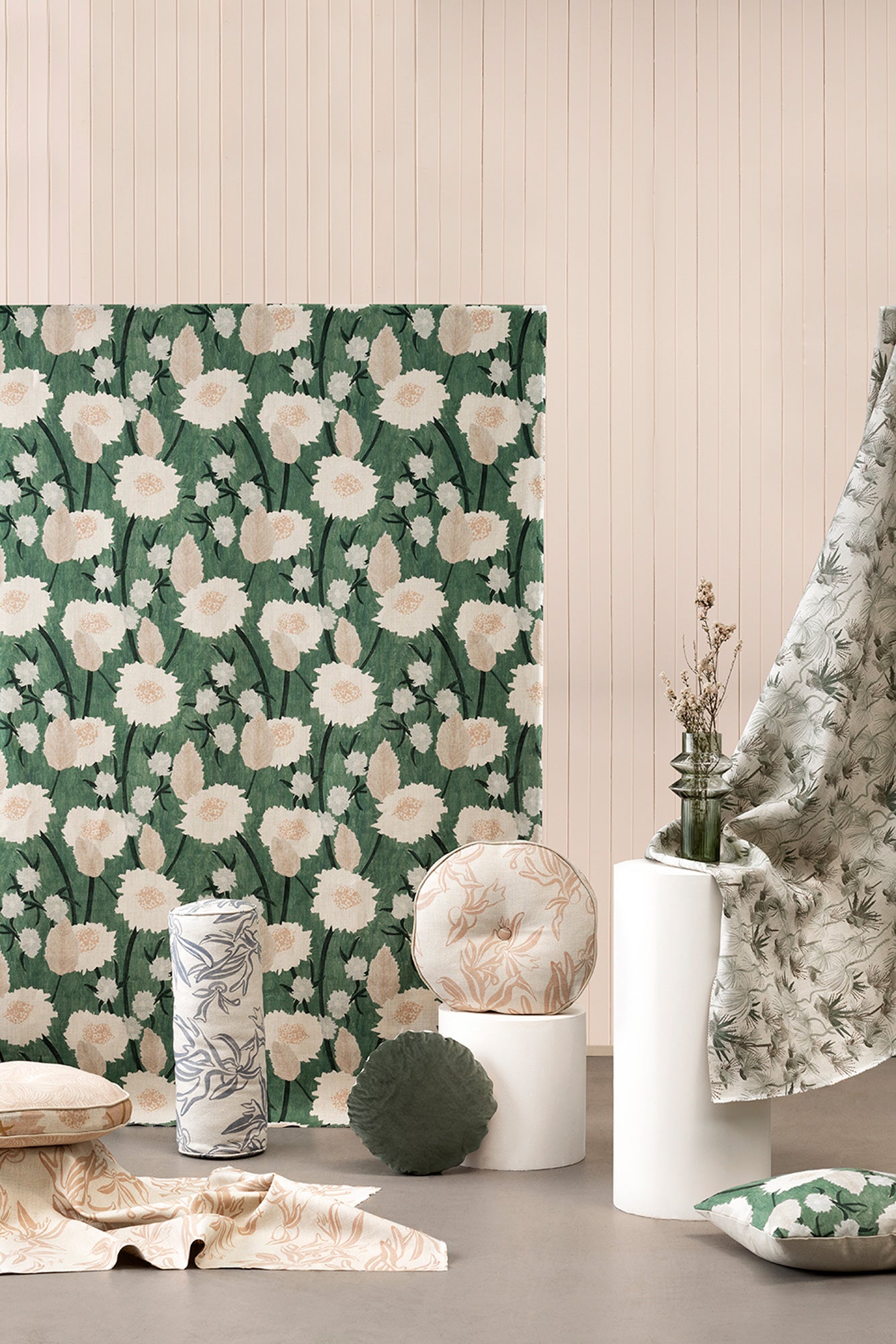 A tableau of pillows, rolls of fabric and draped fabric yardage all stand in front of a light pink wall.