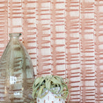 A vase and potted plant stand in front of a wall papered in an undulating ribbon pattern in copper.