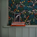 A stack of books leans against a wall papered in a small-scale floral print in blue, green, orange and black.