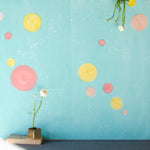 A vase of flowers stands in front of a wall papered in a paint splatter print in multicolor pastels on a turquoise field.