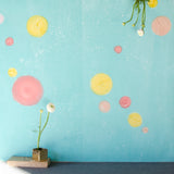 A vase of flowers stands in front of a wall papered in a paint splatter print in multicolor pastels on a turquoise field.