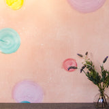 A vase of flowers stands in front of a wall papered in a paint splatter print in multicolor pastels on a light pink field.