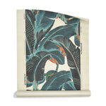 A roll of banana leaf pattern grasscloth wallpaper featuring large tropical leaves in dark charcoal gray, muted teal, and soft orange on an off-white background.