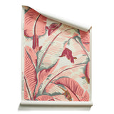 A roll of banana leaf pattern wallpaper featuring large tropical leaves in terracotta pink, cream, and muted gray-green tones on a light cream background.