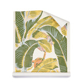 A roll of wallpaper featuring a bold tropical banana leaf pattern in olive green, mustard yellow, and soft peach on a cream background, shown partially unrolled against a clean white studio backdrop.